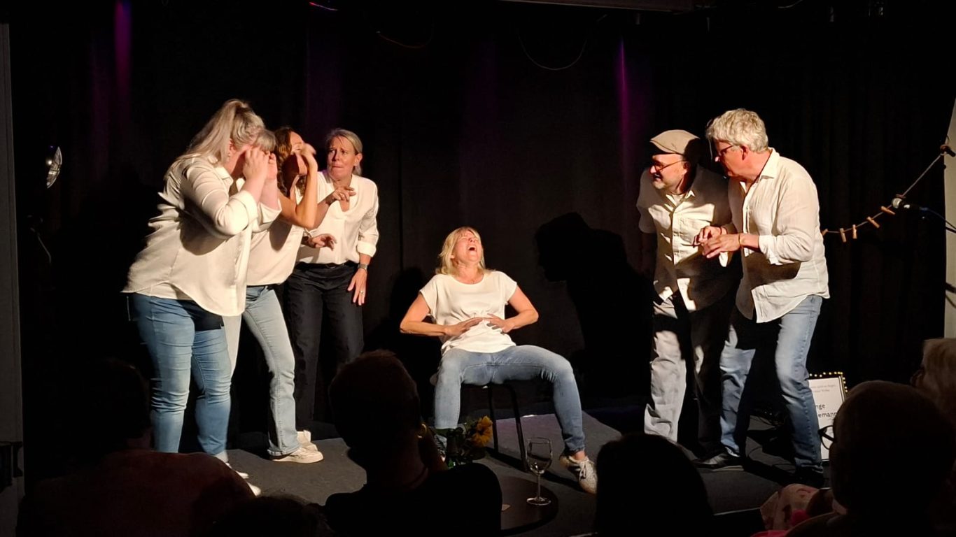 Szenendarstellung: Die Geburt von Inge Eine Schauspielerin sitzt auf einem Stuhl und stellt die Geburt dar, umringt von aufgeregt agierenden Ensemble-Mitgliedern.
