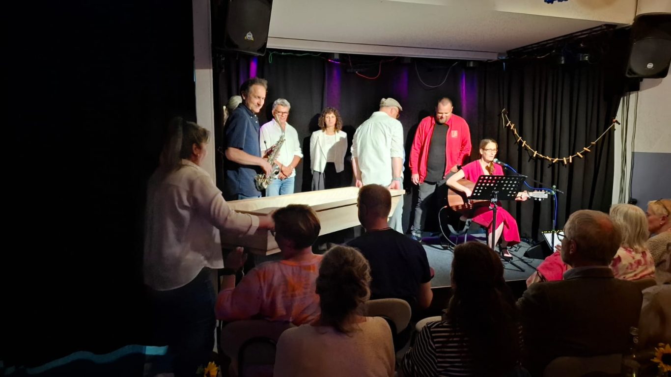 Einzug des Sargs auf die Bühne | Spontanika in Stormarn Ein helfendes Team bringt einen hellen Holzsarg auf die Bühne im Kultavida, während Musiker und Darsteller bereitstehen.
