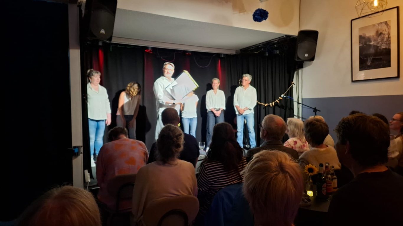 Präsentation der ausgesuchten Totenanzeige auf der Bühne Die Gruppe Spontanika steht auf der Bühne im Kultavida und schaut zu einem Mitglied, das ein Bild auf der Staffelei präsentiert.