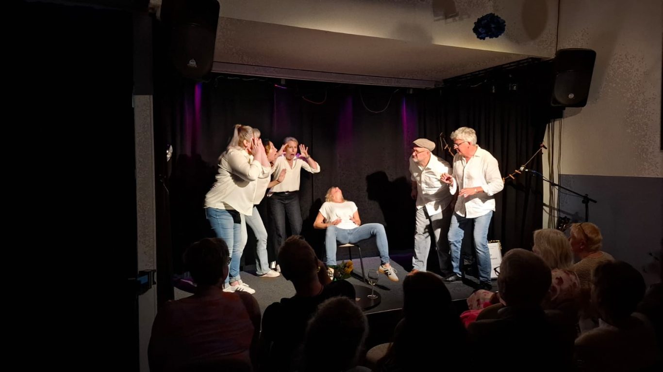 Szenendarstellung: Die Geburt von Inge Eine Schauspielerin sitzt auf einem Stuhl und stellt die Geburt dar, umringt von aufgeregt agierenden Ensemble-Mitgliedern.