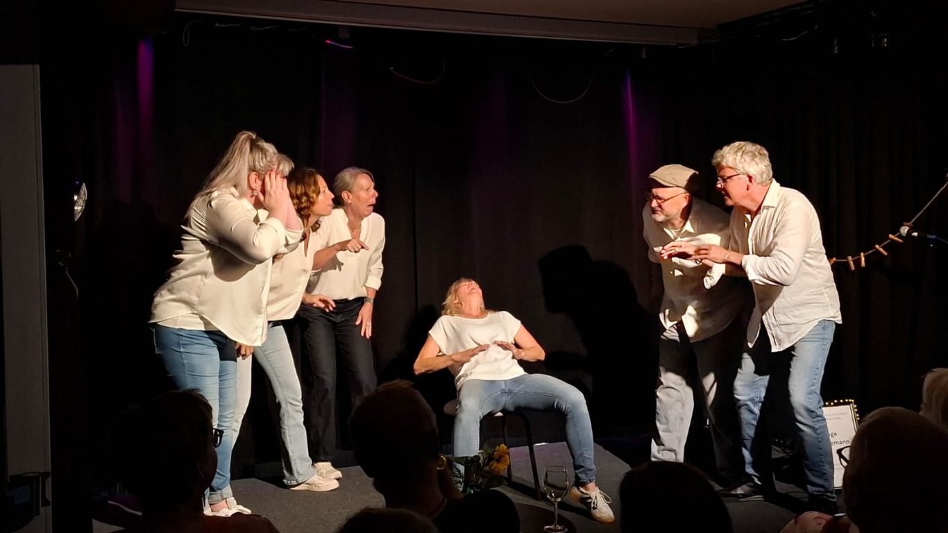 Szenendarstellung: Die Geburt von Inge Eine Schauspielerin sitzt auf einem Stuhl und stellt die Geburt dar, umringt von aufgeregt agierenden Ensemble-Mitgliedern.