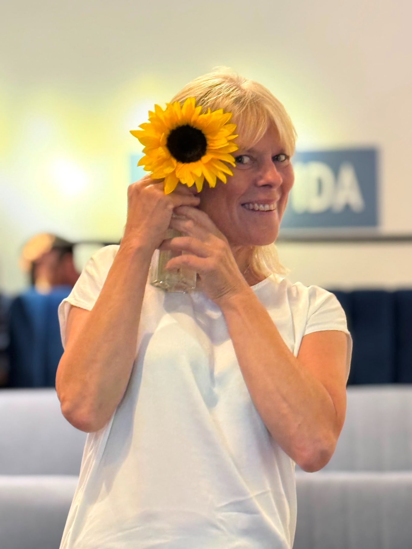 Edith mit Sonnenblume bei „Das letzte Lachen“ im Kultavida Porträt von Edith, einer lächelnden Frau im weißen Shirt, die sich eine Sonnenblume vor das Gesicht hält.