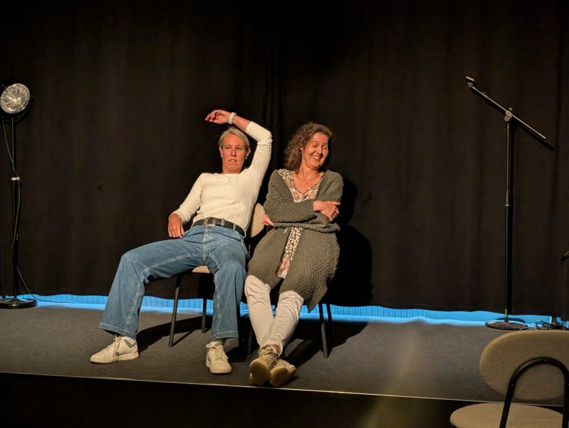 Spontanika-Impro-Szene-Bank Zwei Personen in einer Improvisationsszene auf der Bühne, eine Person gestikuliert mit erhobenem Arm.