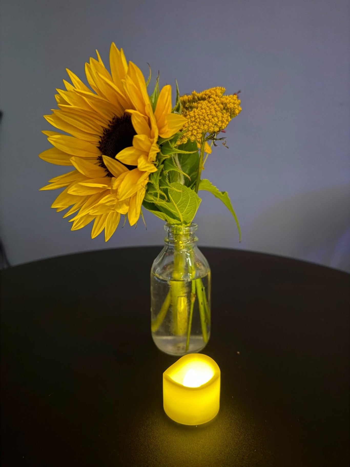 Blumenschmuck und Kerze bei „Das letzte Lachen“ im Kultavida Eine kleine Vase mit einer Sonnenblume und gelben Blumen sowie eine brennende LED-Kerze auf einem Tisch im Kultavida bei „Das letzte Lachen“