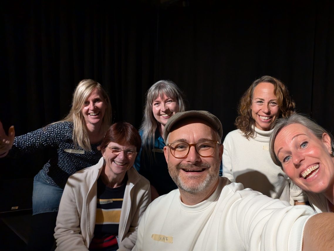Neustart nach der Sommerpause: Improwerkstatt in Ahrensburg im Kultavida Group selfie of the Spontanika improv theater group, smiling brightly together on stage in front of a dark curtain.