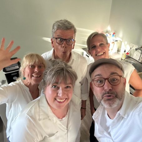 improtheater-spontanika-ensemble-ahrensburg-stormarn Das fünfköpfige Ensemble der Improtheater-Gruppe Spontanika aus Ahrensburg lächelt gut gelaunt in weißen Outfits für ein Backstage-Selfie.