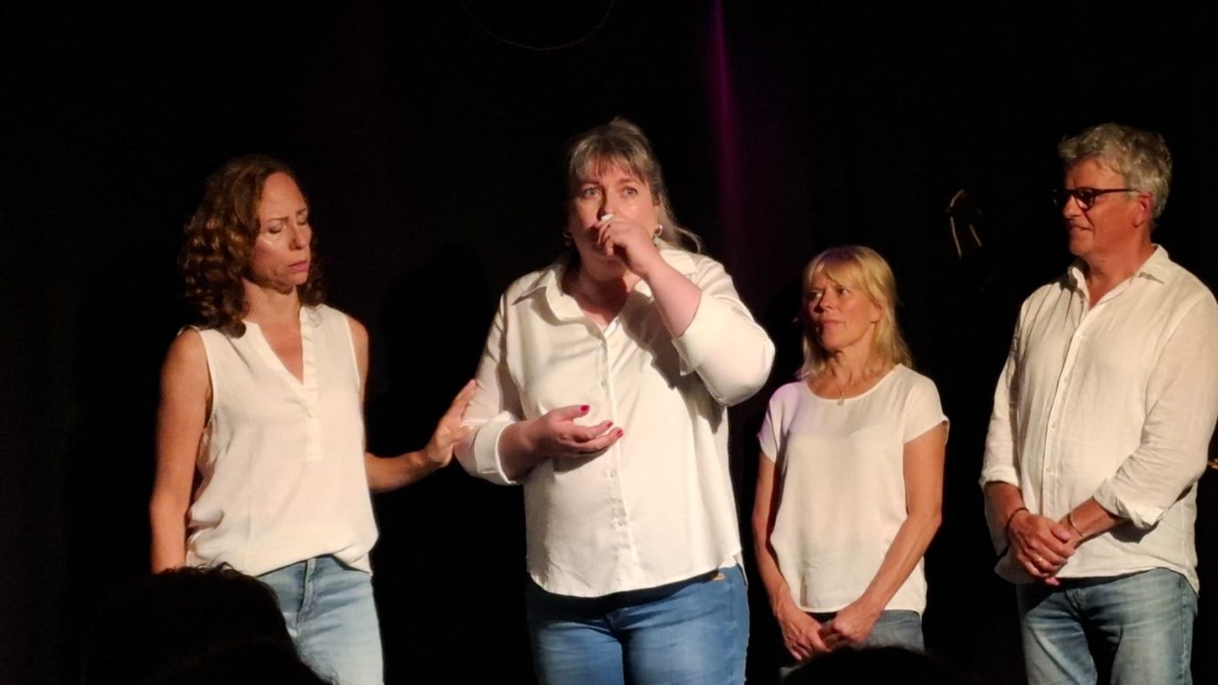 Fokussierte Szene auf der Bühne Vier Ensemble-Mitglieder von Spontanika stehen nebeneinander auf der Bühne, während eine Schauspielerin konzentriert spricht.