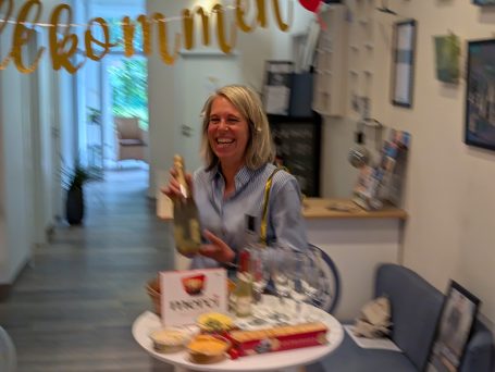 Willkommen, Tanja! Tanja, die neue Mitspielerin der Improgruppe, hält strahlend eine Flasche Sekt zur Begrüßung in der Hand, umgeben von einem Stehtisch