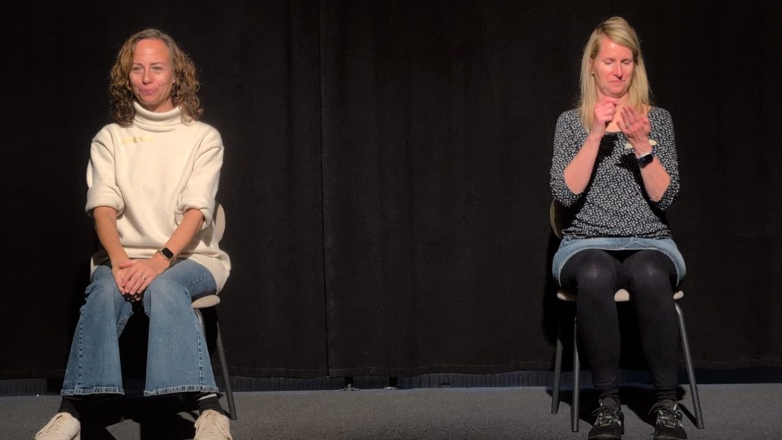 Improwerkstatt Ahrensburg – Szene aus dem Improtheater-Spiel Briefwechsel Zwei Frauen beim Improtheater-Workshop in Ahrensburg auf Stühlen während der Übung Briefwechsel auf der Bühne.