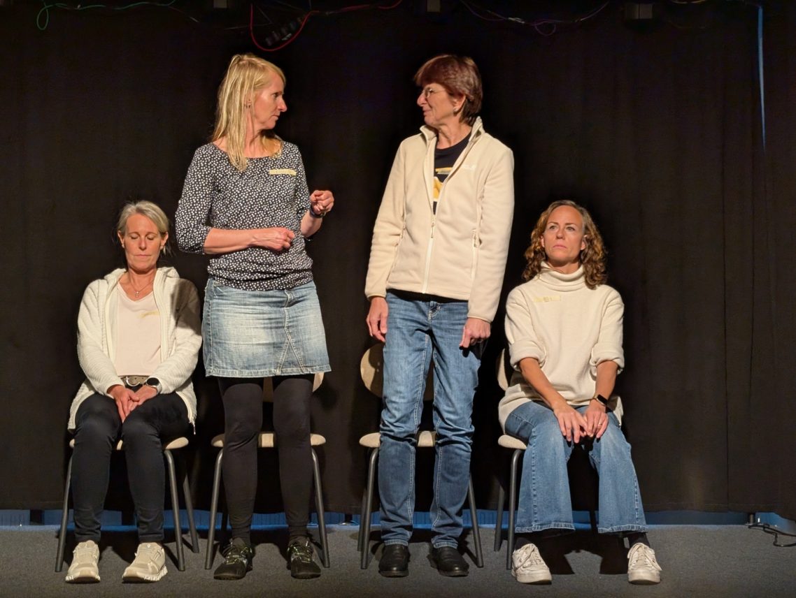 Improwerkstatt nach der Sommerpause – Toaster-Spiel Vier Personen bei einer Improtheater-Übung auf der Bühne: Zwei Personen stehen in der Mitte und interagieren miteinander, während links und